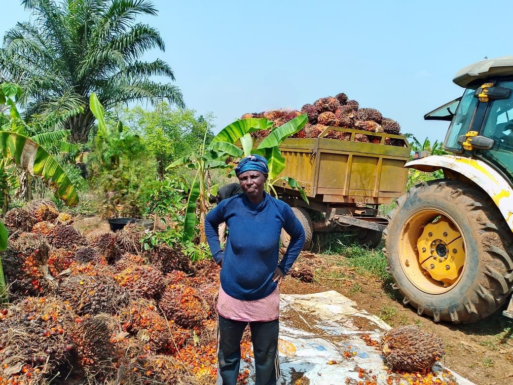 A smallholder farmer standing beside her harvested fresh fruit bunches, with a loaded trailer behind her